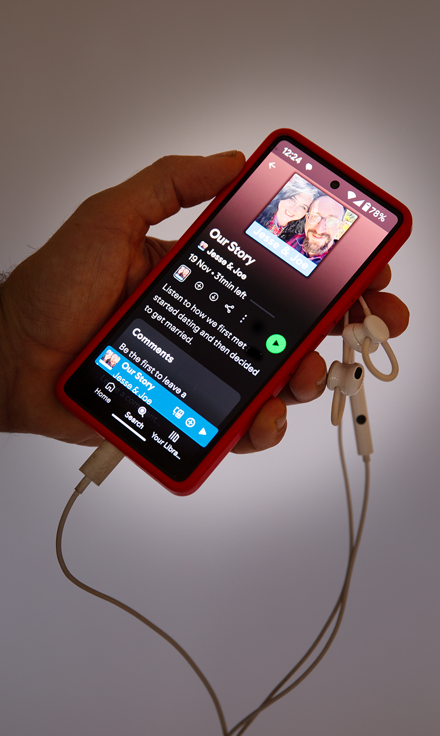 Photograph of a hand holding headphones and a mobile phone displaying a private wedding podcast about Jesse and Joe.